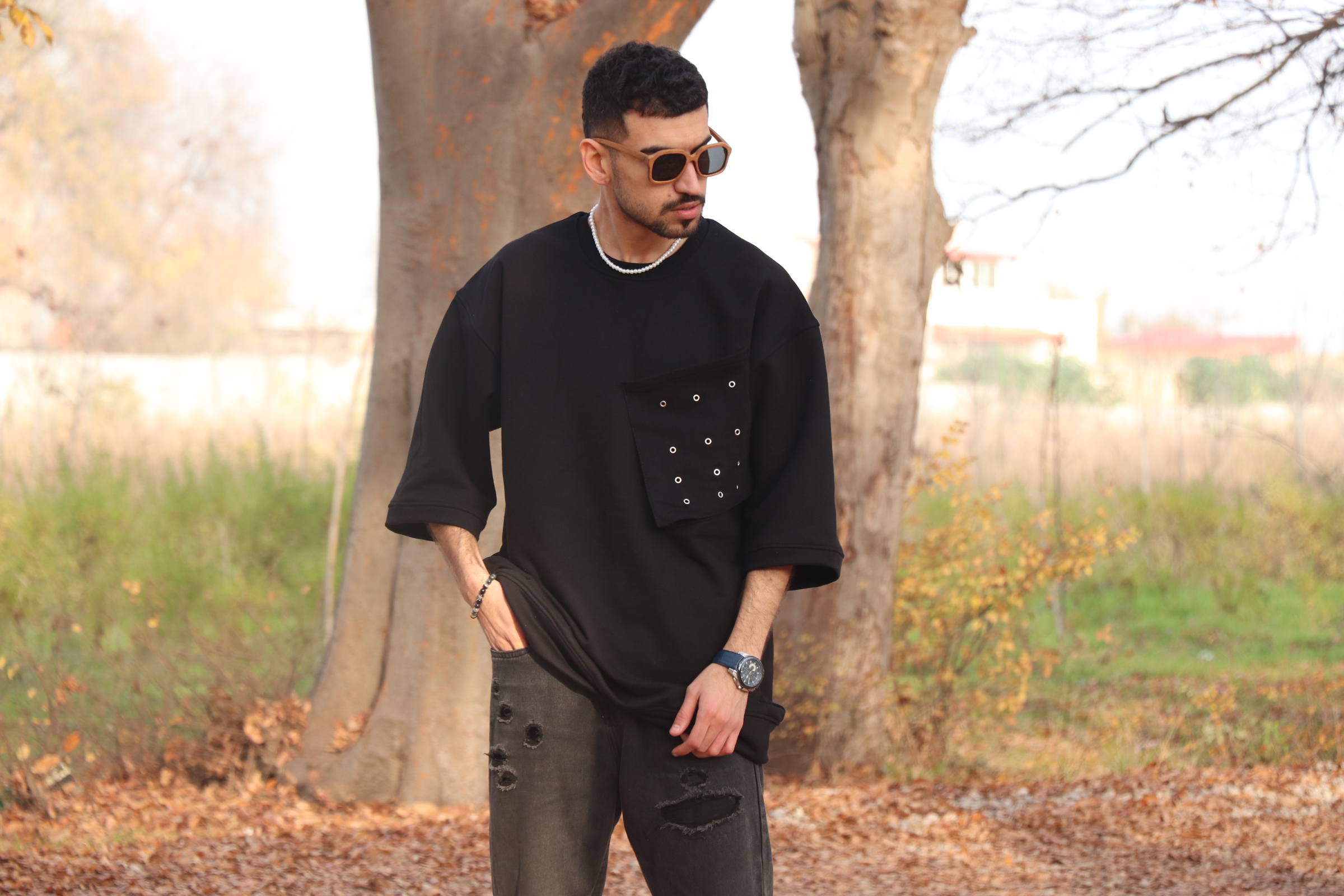 Matin, a young man with short, curly black hair and a well-groomed beard stands outdoors in a park-like setting during autumn. He is wearing a black oversized t-shirt with a large pocket on the left side, decorated with multiple silver eyelets, along with distressed black jeans featuring several ripped sections. He accessorizes with a silver chain necklace, a blue wristwatch, a black bracelet, and wooden-framed sunglasses with brown-tinted lenses. His expression is neutral as he gazes to the side. The background consists of tall, leafless trees with textured bark, fallen leaves on the ground, and a blurred view of houses and greenery in the distance.