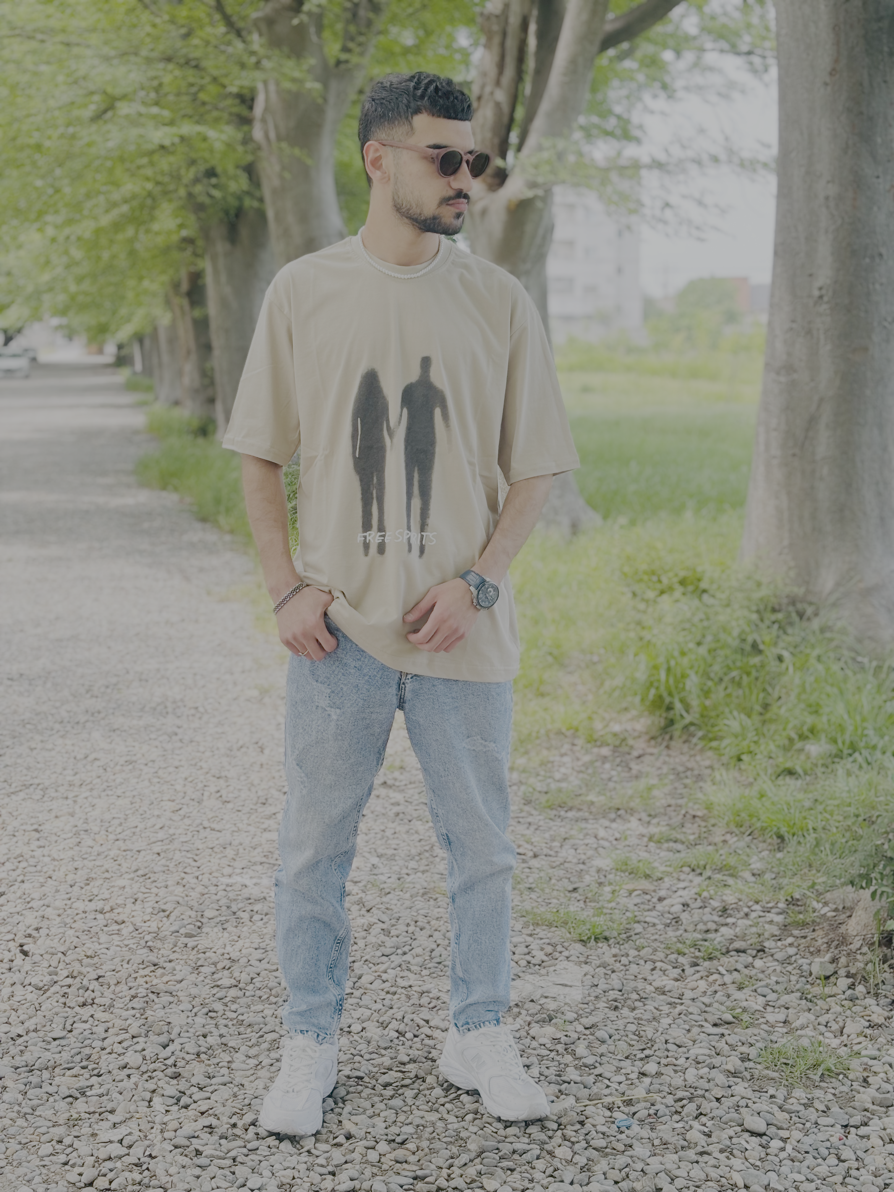 Matin, a young man with short, curly black hair and a well-groomed beard, stands on a gravel path lined with tall trees and lush greenery. He is wearing a beige oversized t-shirt featuring a black stencil-style graphic of a man and woman holding hands, with the words ‘FREE SPIRITS’ written underneath. He pairs the shirt with light blue denim jeans and white sneakers. He accessorizes with a silver chain necklace, a wristwatch, a bracelet, and round brown sunglasses with a thin frame. His posture is relaxed, with one hand on his shirt and the other resting on his pocket as he gazes off to the side. In the background, a few buildings are visible beyond the green landscape, adding depth to the serene outdoor setting.