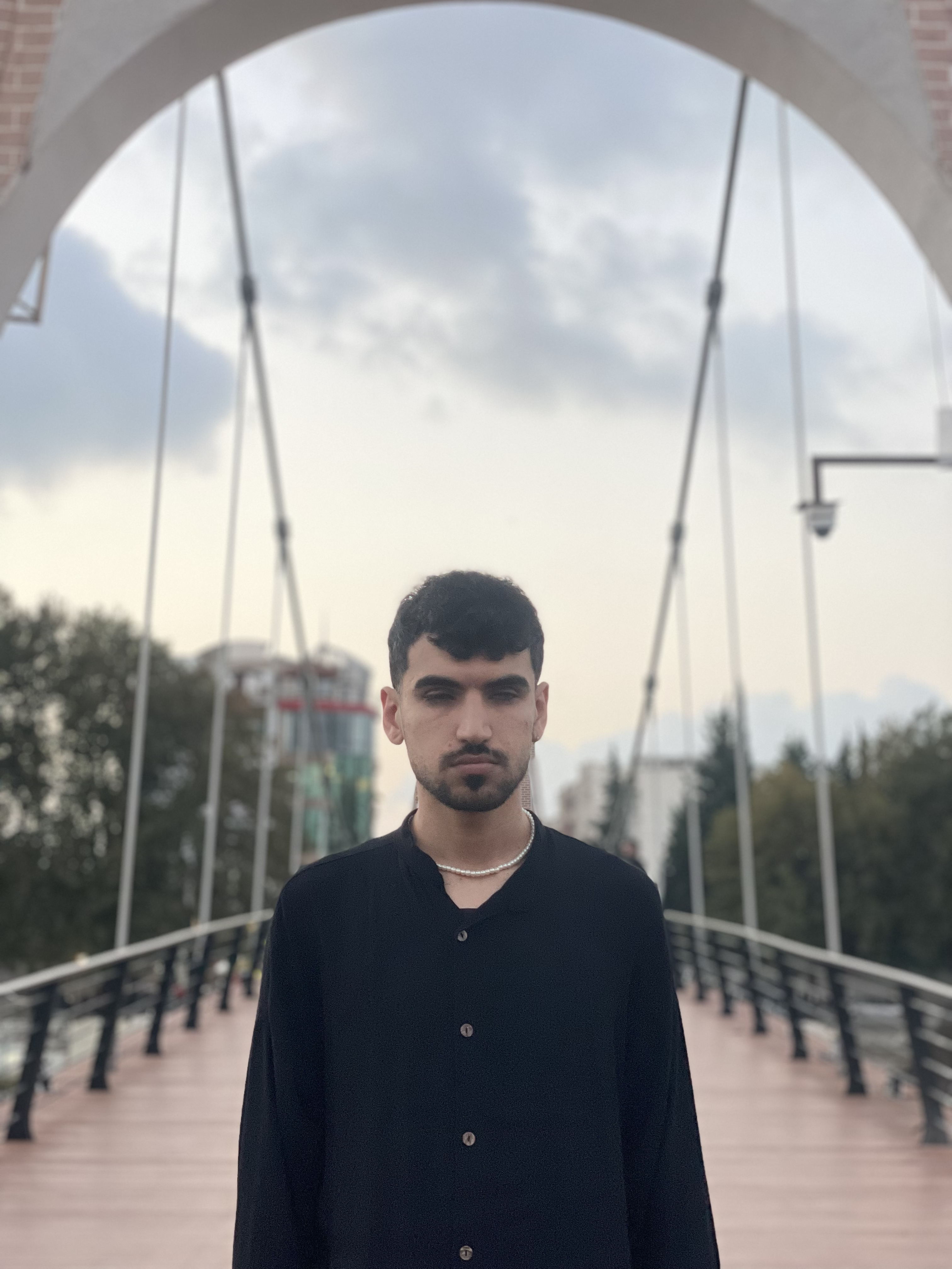 Matin, a young blind man with short, curly black hair and a well-groomed beard, stands on a modern suspension bridge with metal cables and an arched entrance framing the sky behind him. He is wearing a black button-up shirt with a relaxed fit and a pearl necklace. His expression is calm and composed. The background features a cityscape with buildings, trees, and a cloudy sky, adding depth to the scene. The soft lighting and slightly blurred background create a moody, introspective atmosphere.