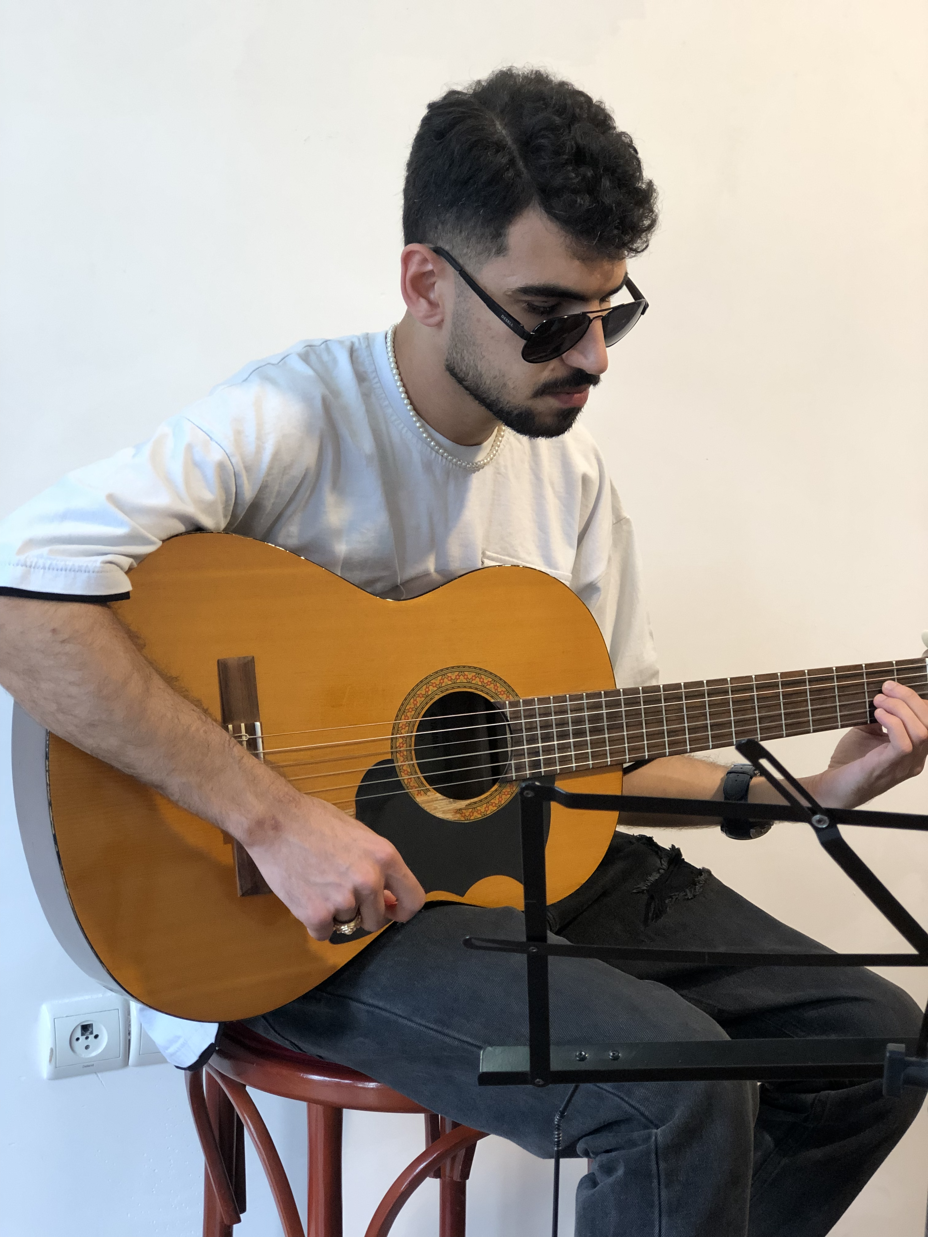 Matin, a young man with short, curly black hair and a well-groomed beard, sits on a wooden stool, playing a classical acoustic guitar. He is wearing a light-colored t-shirt, black distressed jeans, a pearl necklace, and dark sunglasses. His fingers are positioned carefully on the fretboard as he plays, with a focused and serene expression. A black music stand is positioned in front of him, though he does not rely on sheet music. The background is minimal, with a white wall and an electrical outlet visible near the floor. The soft lighting and neutral tones create an intimate, calm atmosphere.